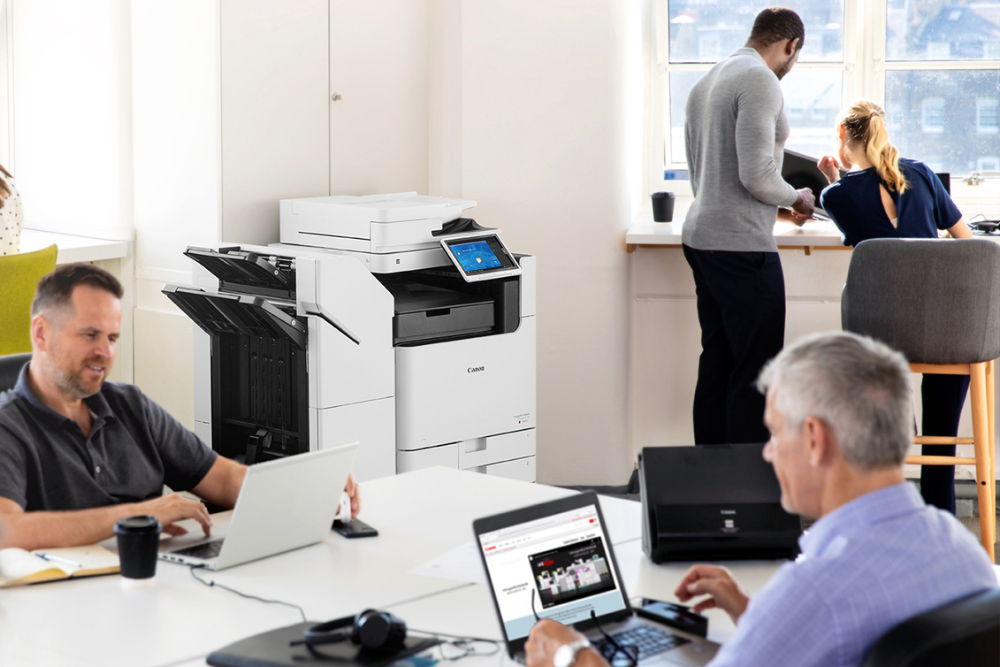 Modern office with employees working on laptops and an office multifunction printer in the background, highlighting its use in a collaborative workspace.