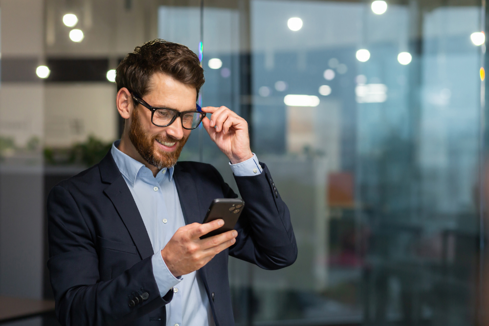 A professional man in a modern office smiling as he uses his smartphone, representing the convenience and accessibility of mobile printing. This image highlights the ability to manage printing tasks directly from mobile devices, enhancing workplace efficiency.