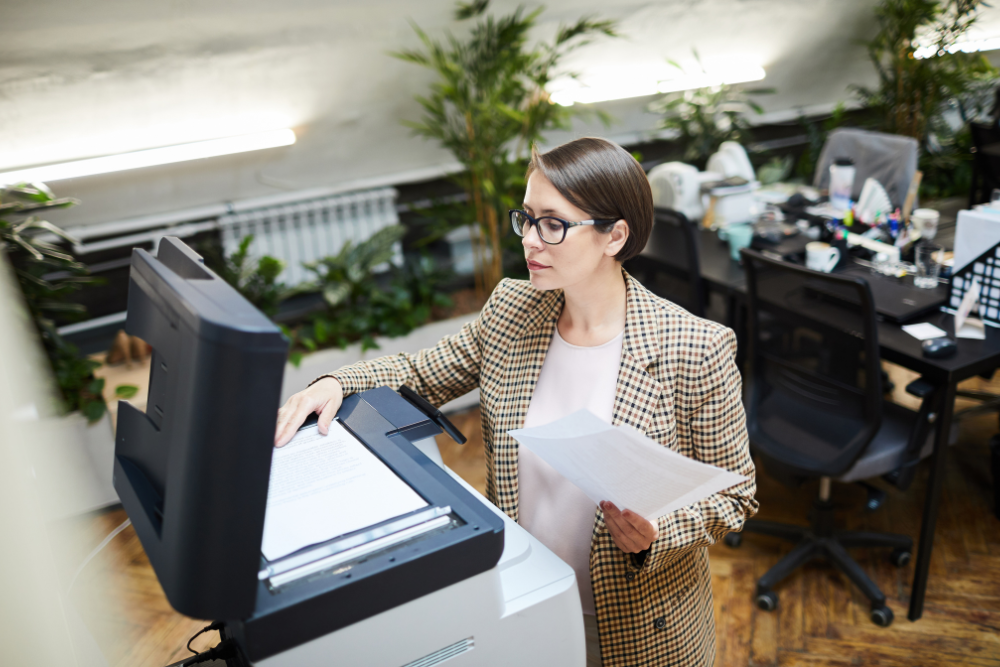 A professional woman in a modern office using an office copier to make copies of documents. This image illustrates the everyday use of office copiers in a business setting, highlighting their importance in document management and productivity.