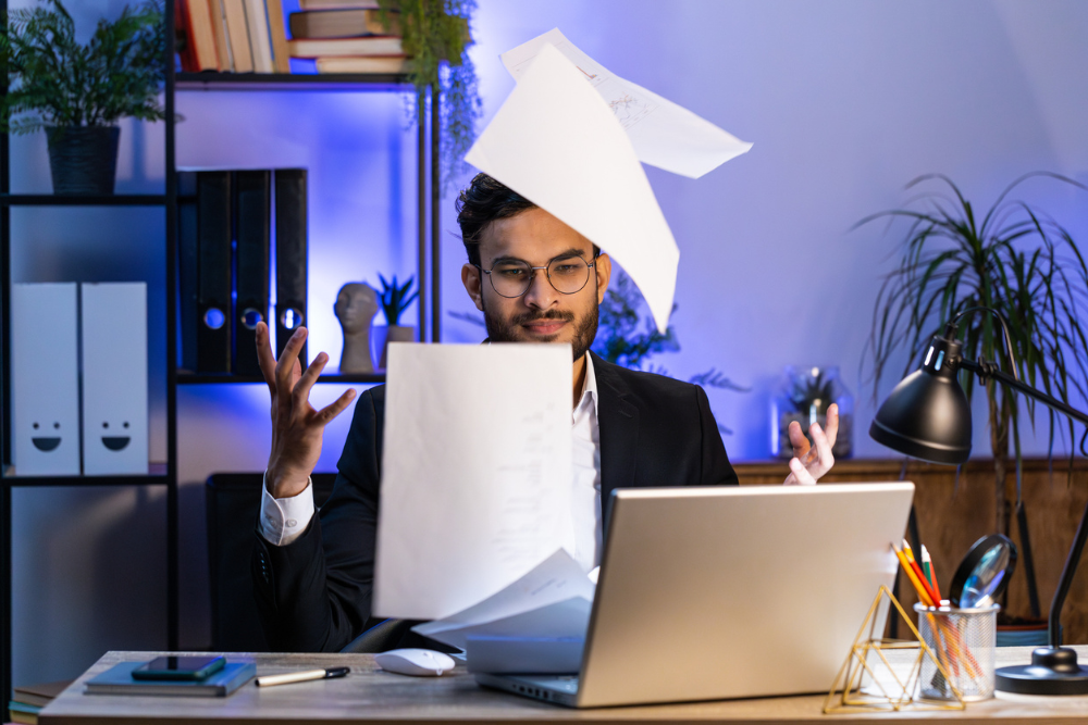 A frustrated office worker tosses papers into the air while sitting at a desk with a laptop, highlighting the need for effective document management system. The background includes shelves with books and plants in a modern office setting.