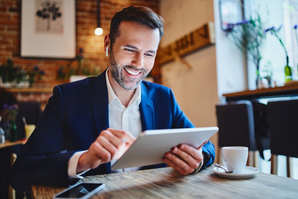 A smiling professional sitting in a modern café, using a tablet while wearing wireless earbuds. The setting highlights seamless communication and productivity, showcasing the benefits of business VoIP solutions for remote and mobile work environments.