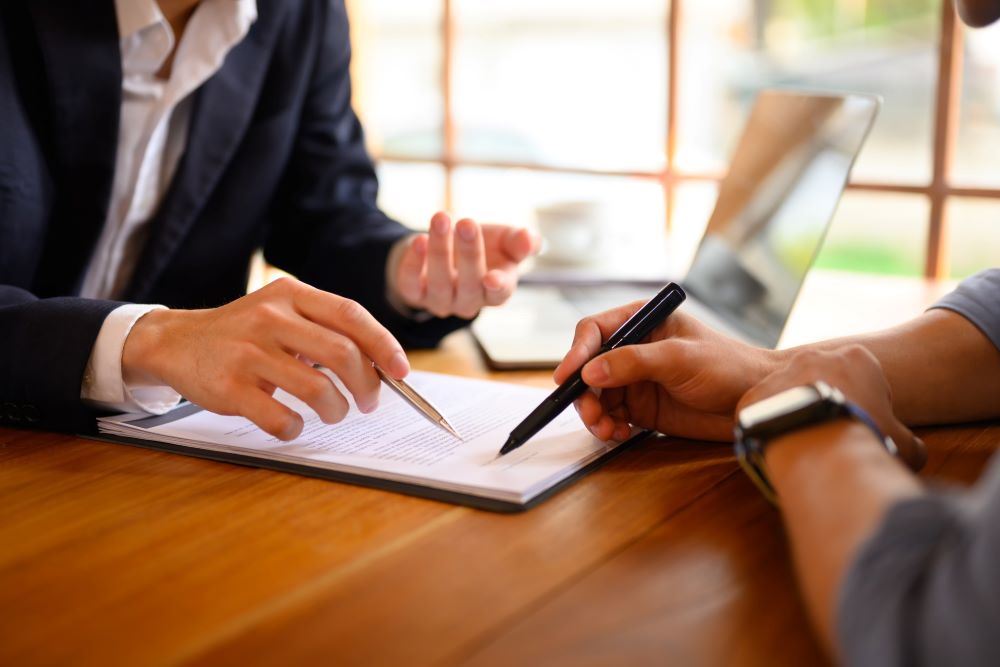 Two business professionals discussing and reviewing documents at a wooden desk, with a focus on an office copier lease agreement. One person is pointing at the paper with a pen, highlighting important details, while the other attentively listens, showcasing a collaborative business setting.