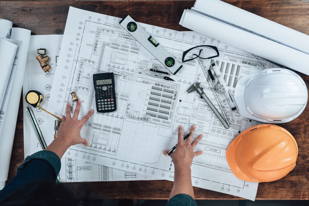 An engineer or architect reviewing blueprints on a desk filled with tools, hard hats, and measuring instruments. The detailed plans highlight the importance of using a wide format printer for producing accurate and high-quality large-scale documents needed for construction and design projects.