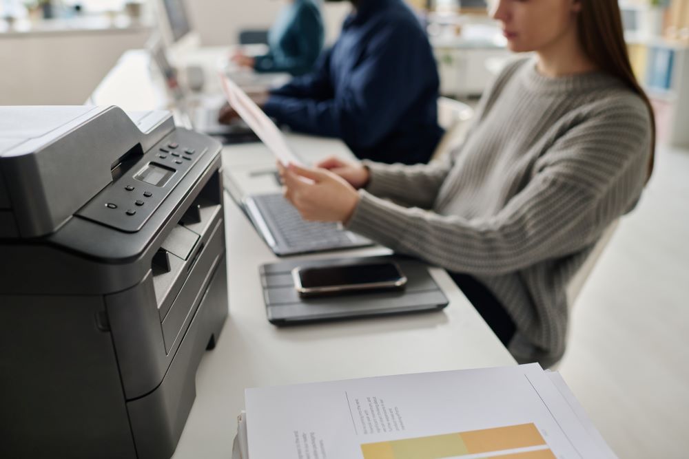 A modern workspace featuring an all-in-one office printer on a desk, with a person reviewing printed documents while working on a laptop nearby.