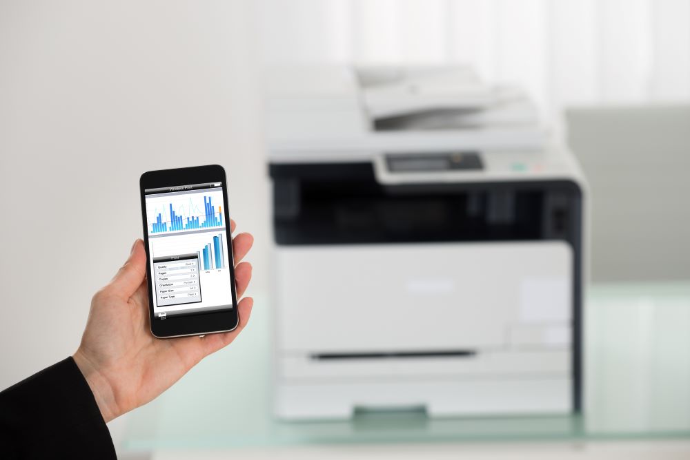 A person holding a smartphone displaying printing options and graphs, with a multifunction printer in the background, showcasing mobile printing capabilities.