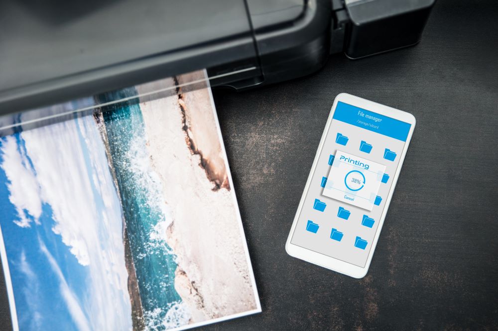 A smartphone displaying a cloud printing progress screen next to a printer producing a high-quality photo of a beach, illustrating wireless and remote printing technology.